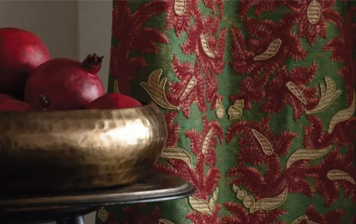 Close-up of Balaustine fabric by Marvic featuring rich red botanical motifs on a green background beside a brass bowl with pomegranates