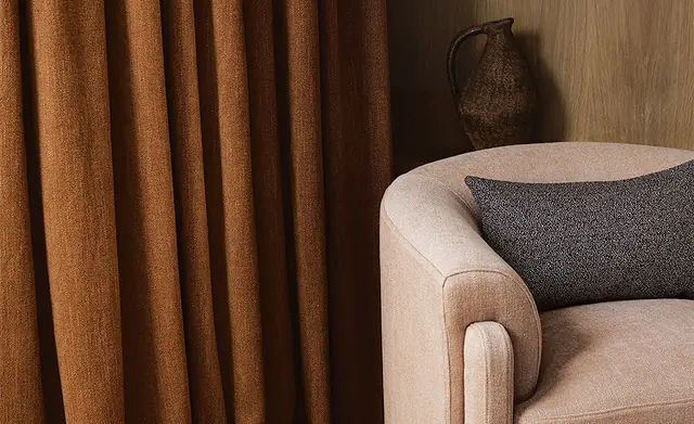 Textured rust curtains beside a cream upholstered chair and woven cushion